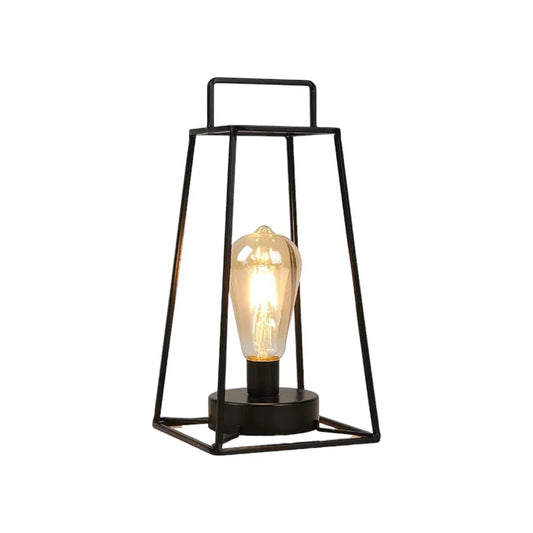 Lifestyle shot of the black cordless geometric metal lantern with a lit Edison bulb on a light wood cabinet beside a potted plant.