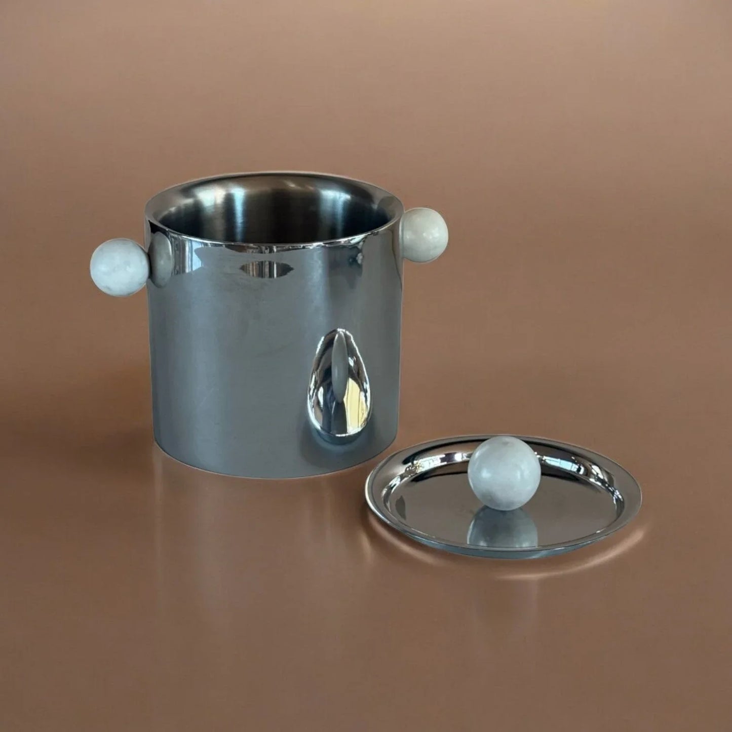 Modern stainless steel ice bucket with marble sphere handles and matching lid on brown surface