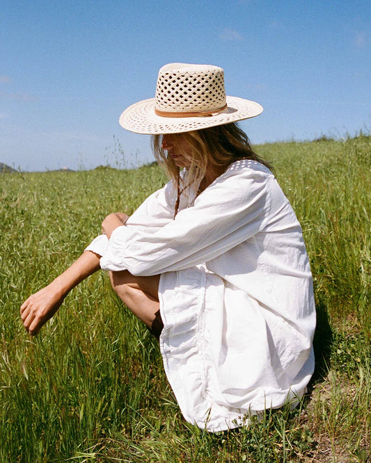 Woman in white dress and woven straw hat sitting in green grassy field under blue sky