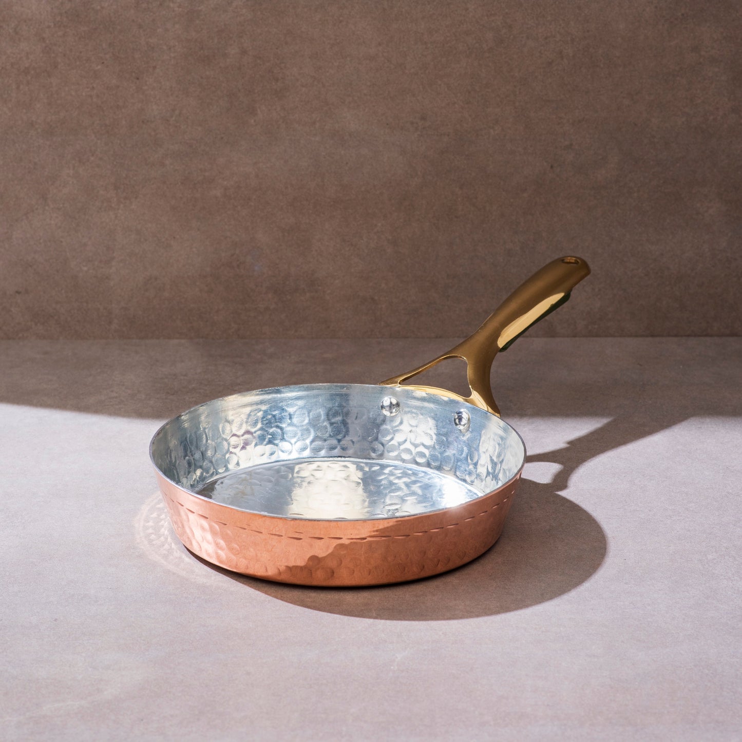 Copper frying pan with hammered exterior and tin lining, features a golden brass handle on a neutral background.