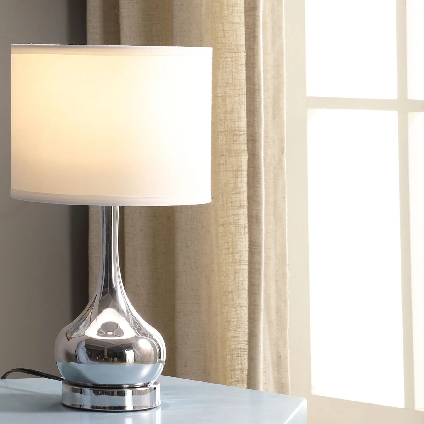 Modern table lamp with chrome base and white drum shade on blue surface near window and beige curtains