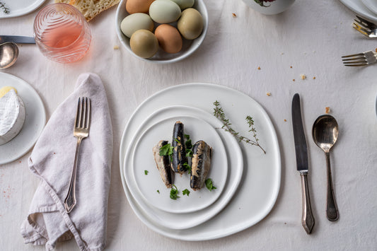 Matte white ripple dinner set with three sardines and herbs, rustic eggs, bread, and silver cutlery on table