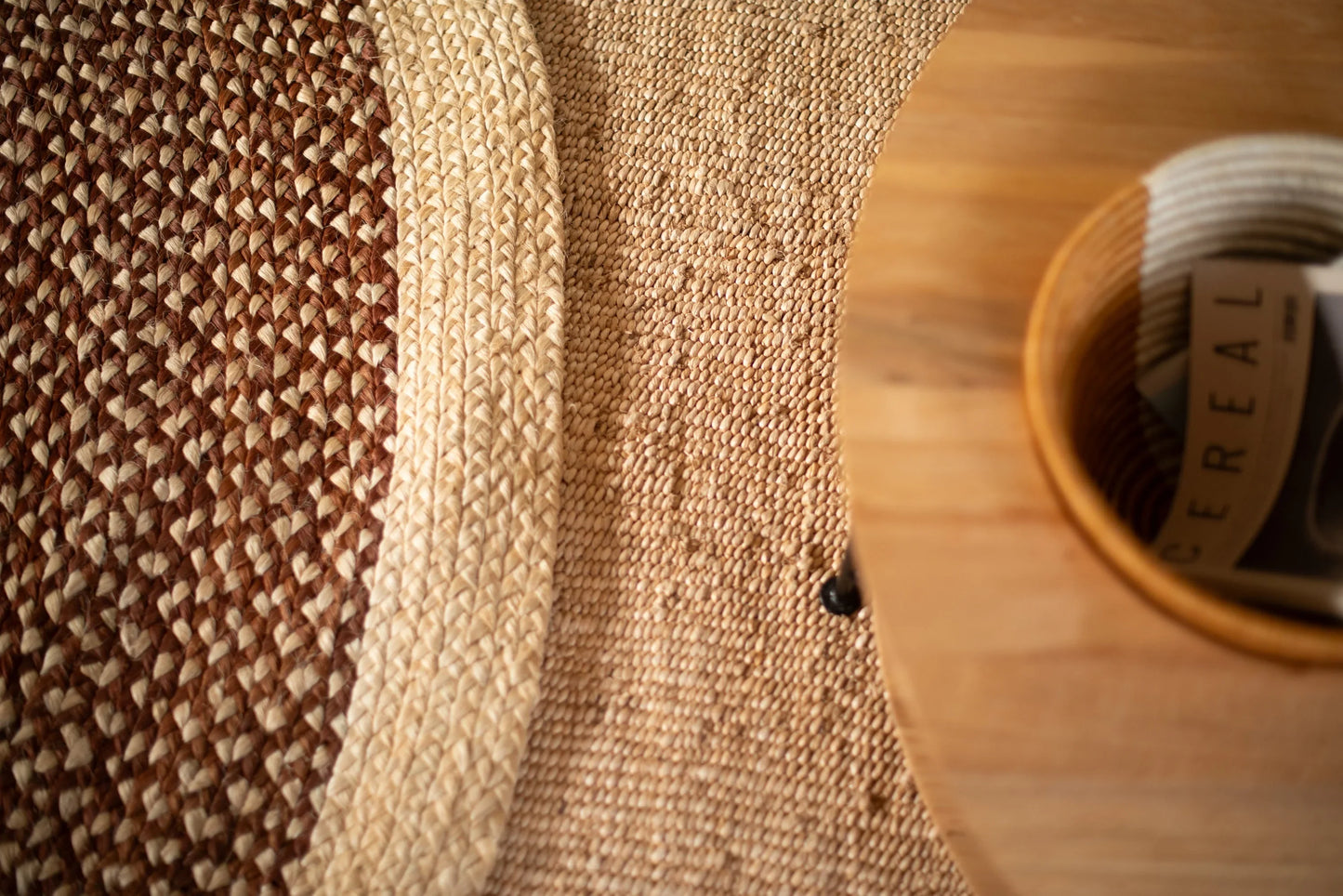 Overhead lifestyle detail of medium round sisal rug red earth variant showing curved cream ring motif separating muted rose brown braided field from natural sisal border with round wood tray and rolled magazine in foreground