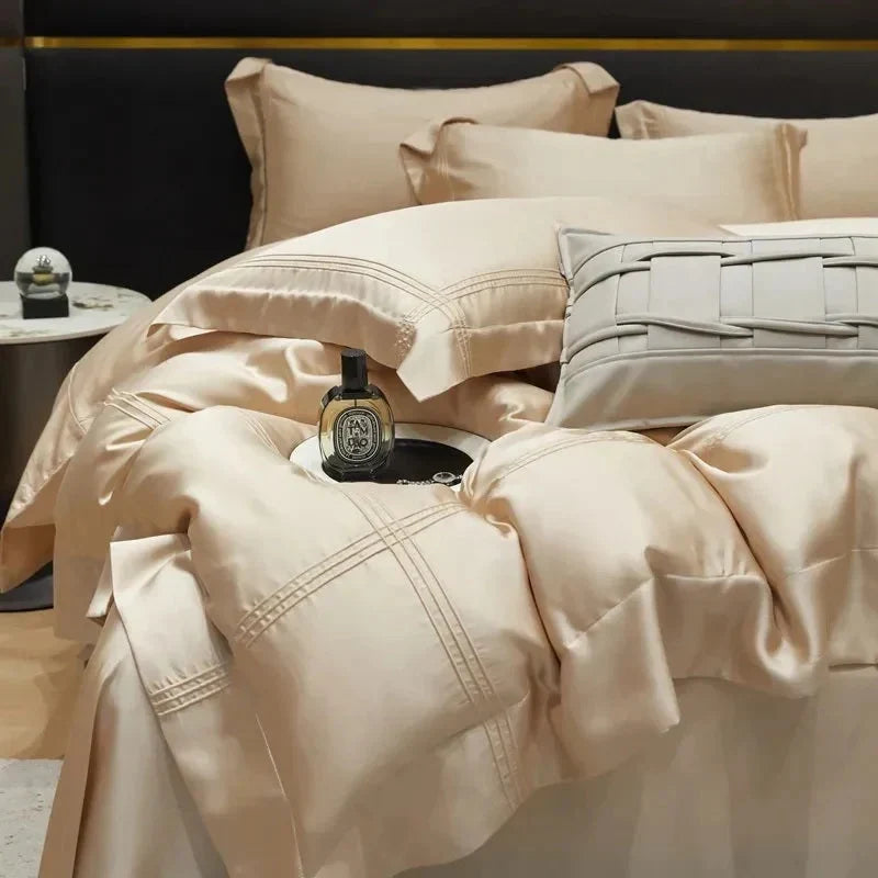 A luxurious Bed & Bath collection display featuring a champagne-toned duvet with refined stitching and textured accent pillows arranged in a modern bedroom setting.