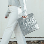 Woman walking, holding a Glacial Gray Woven Laptop Clutch, showcasing the woven texture and cut-out handles.