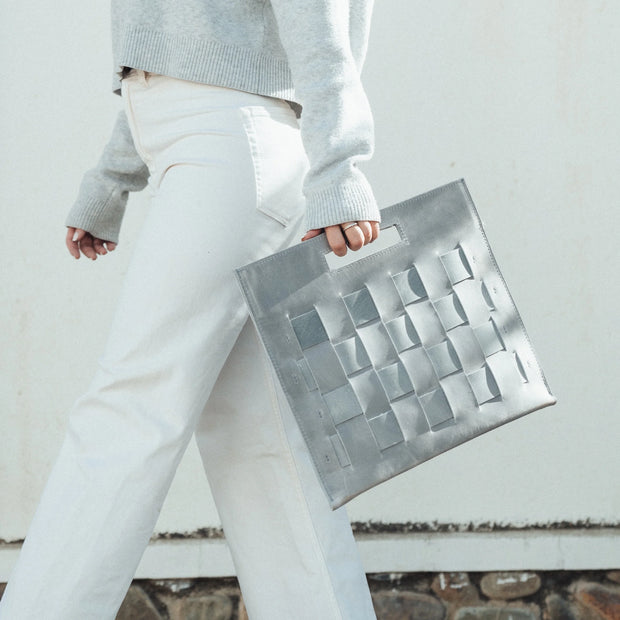 Woman walking, holding a Glacial Gray Woven Laptop Clutch, showcasing the woven texture and cut-out handles.