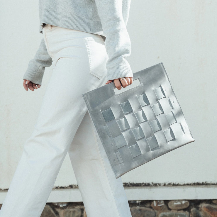 Woman walking, holding a Glacial Gray Woven Laptop Clutch, showcasing the woven texture and cut-out handles.