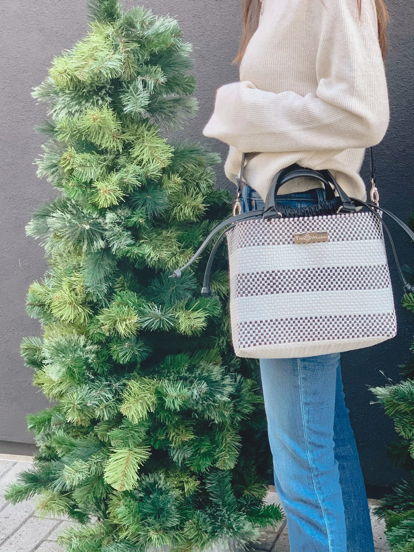 Woman wearing beige sweater and jeans holding a striped woven handbag next to a green pine tree