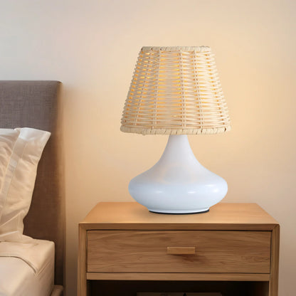 White ceramic table lamp with woven rattan shade on wooden bedside table next to bed