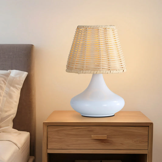 White ceramic table lamp with woven rattan shade on wooden bedside table next to bed