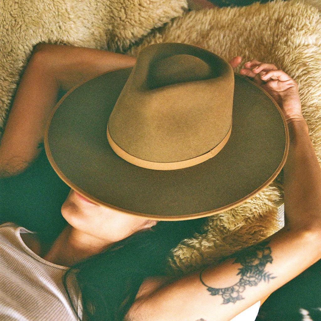 Woman lying on a textured rug wearing a wide-brimmed brown hat and white tank top, showcasing a floral tattoo