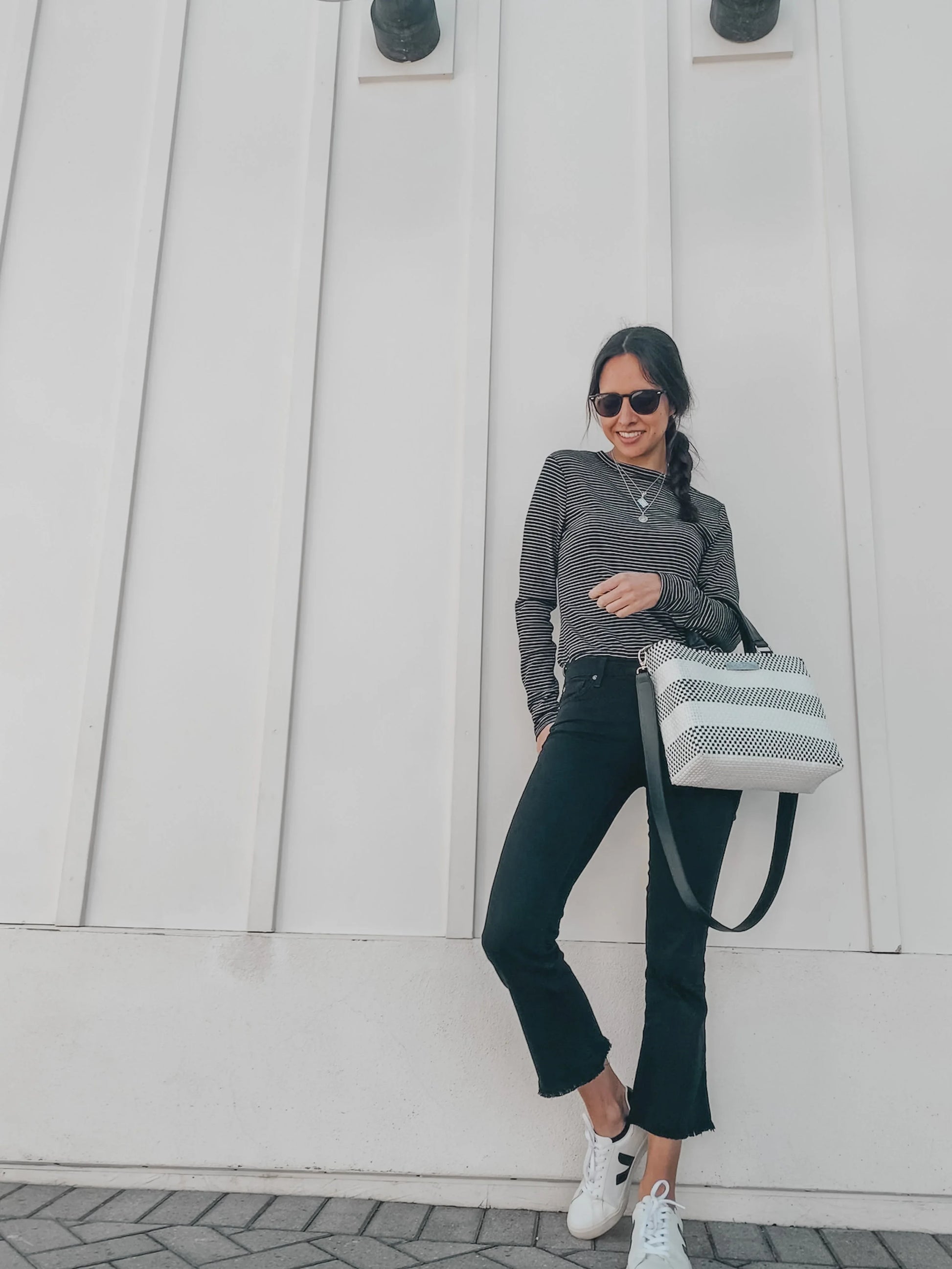Woman wearing striped long-sleeve top, black cropped jeans, white sneakers, sunglasses, and holding black and white striped shoulder bag