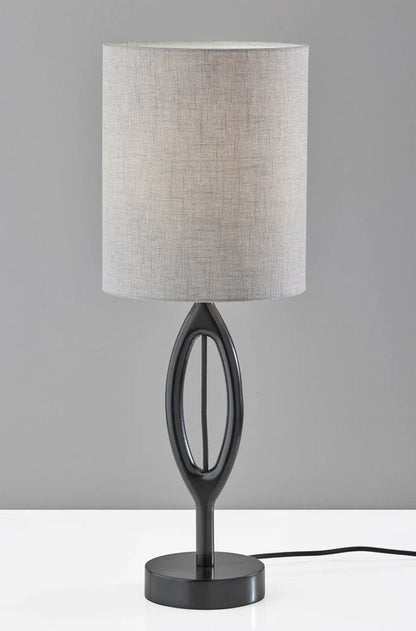 A sculpted black wood table lamp featuring a gray drum shade against a dark grey and white studio background.