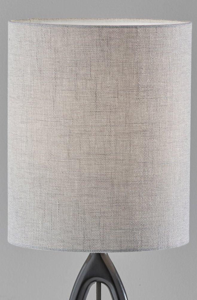 Illuminated gray drum fabric shade of a black wood table lamp showing a warm glow through the textured linen weave against a dark grey background.