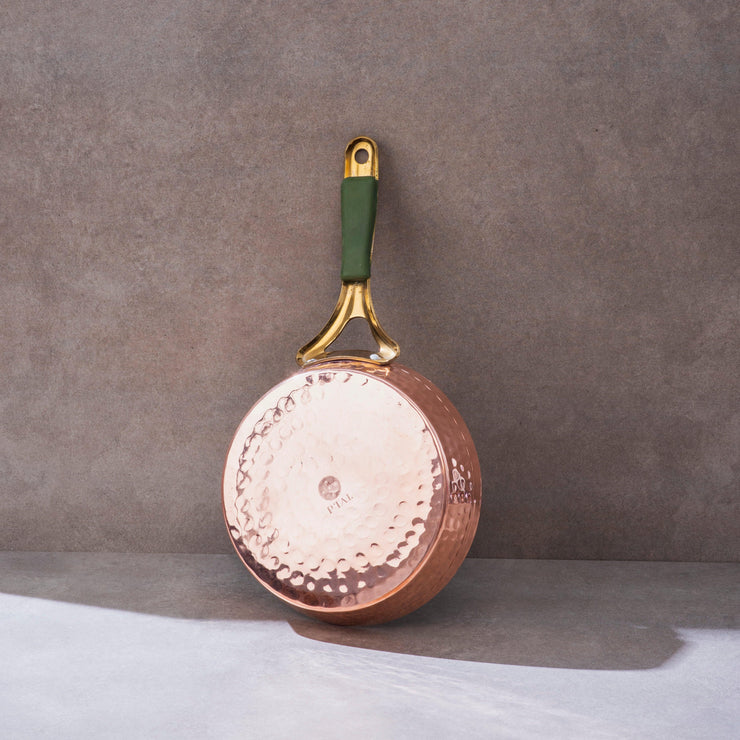 Hammered copper frypan with a shiny brass handle, featuring a green grip, on a neutral background.