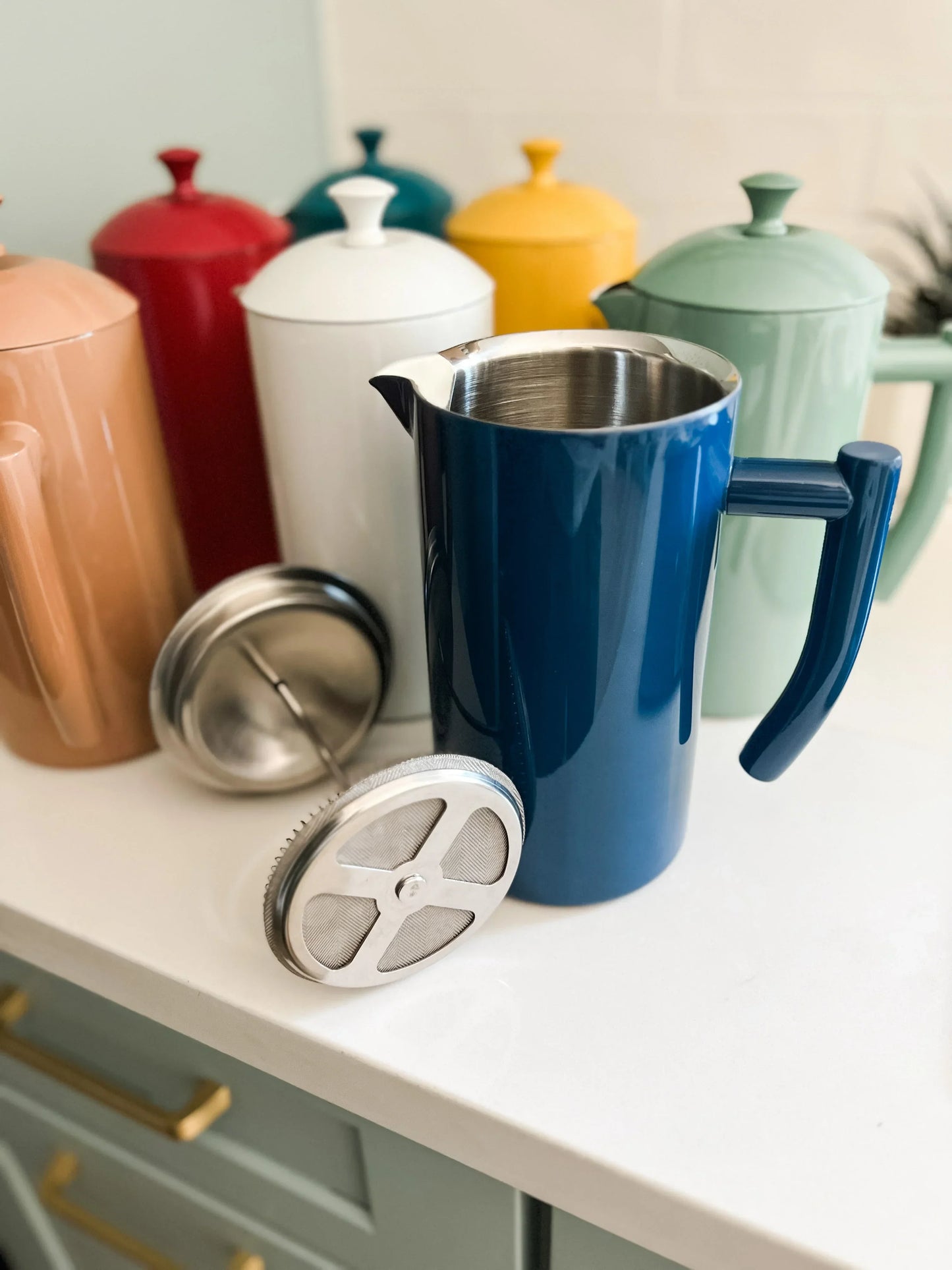 Blue stainless steel French press coffee maker with colorful ceramic coffee pots on kitchen counter