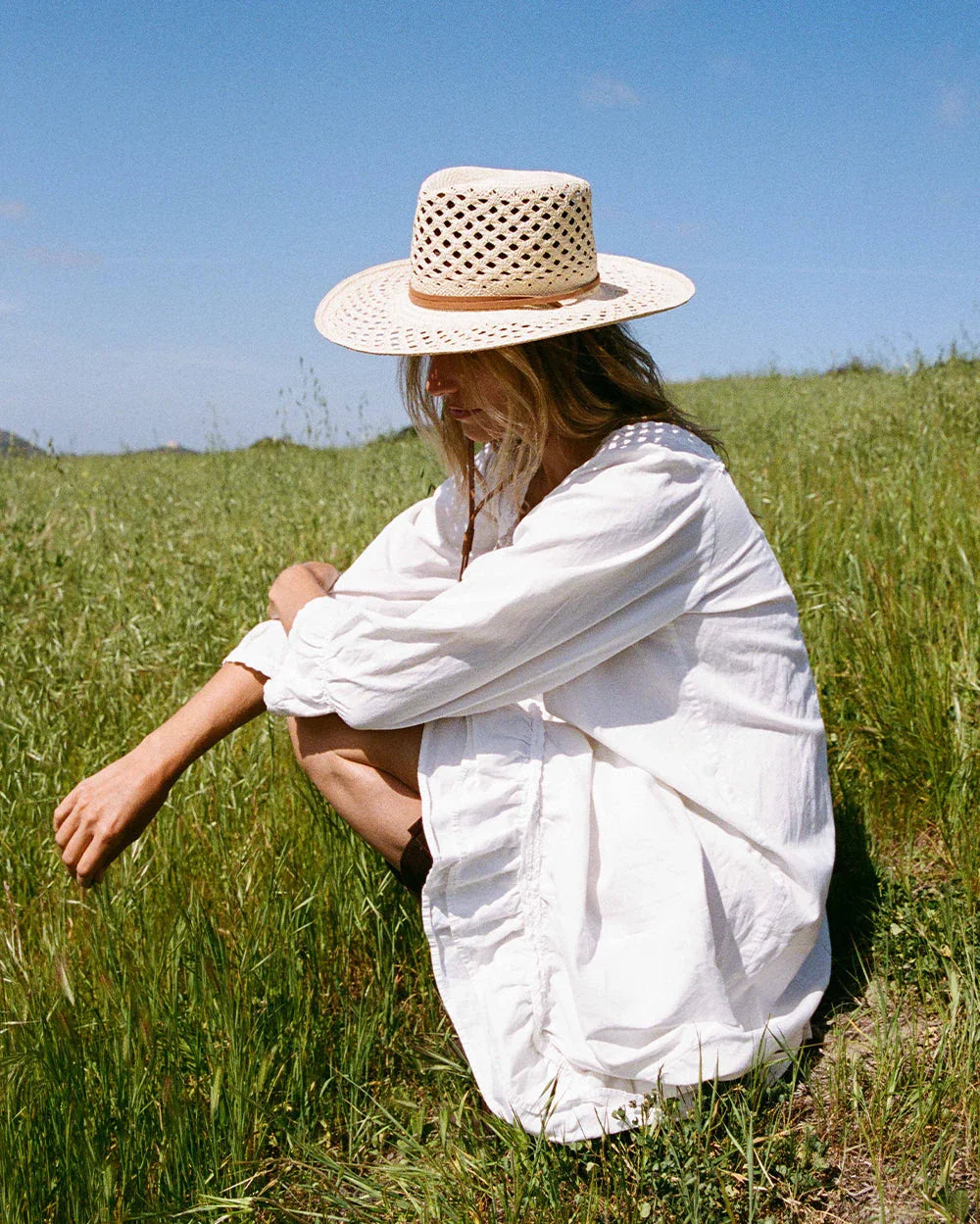 Woman in white dress and woven straw hat sitting in green grassy field under blue sky