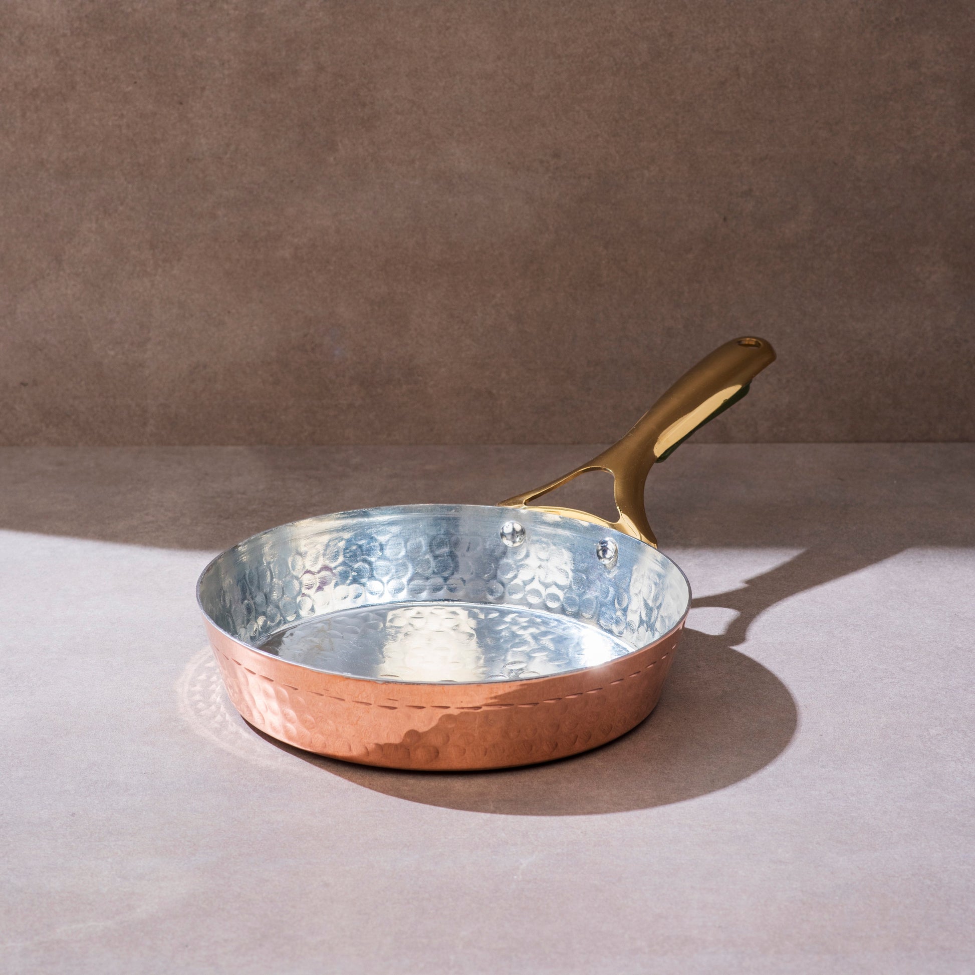 Copper frying pan with hammered exterior and tin lining, features a golden brass handle on a neutral background.