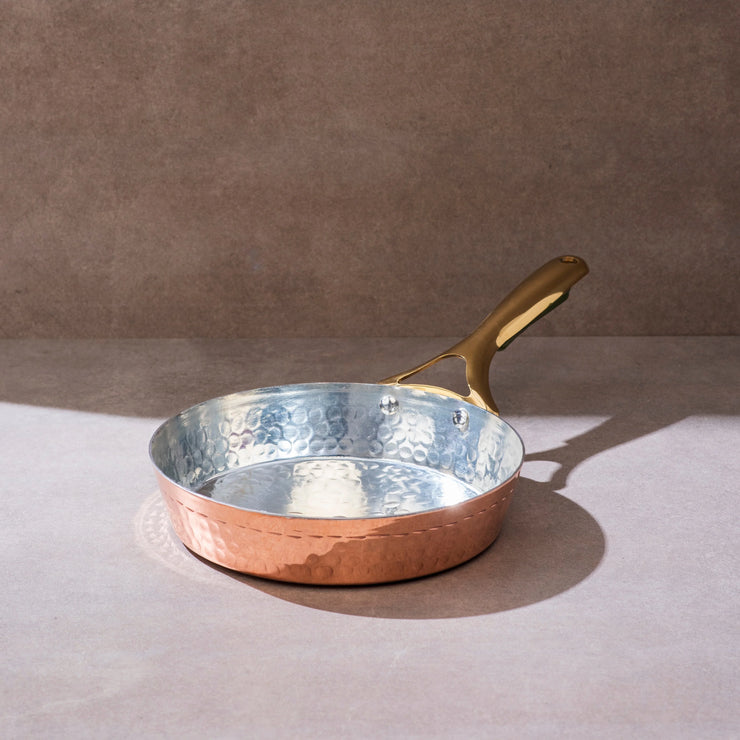 Copper frying pan with hammered exterior and tin lining, features a golden brass handle on a neutral background.