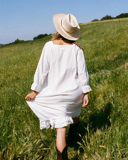 Woman in white dress and straw hat walking through green grassy field with blue sky