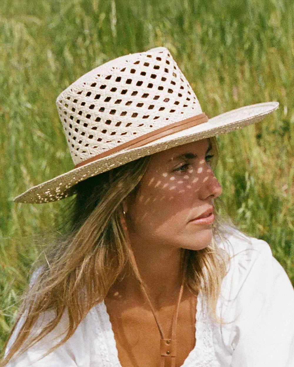 Woman wearing a white perforated straw hat with a tan band, sitting outdoors in green grass, dressed in a white top
