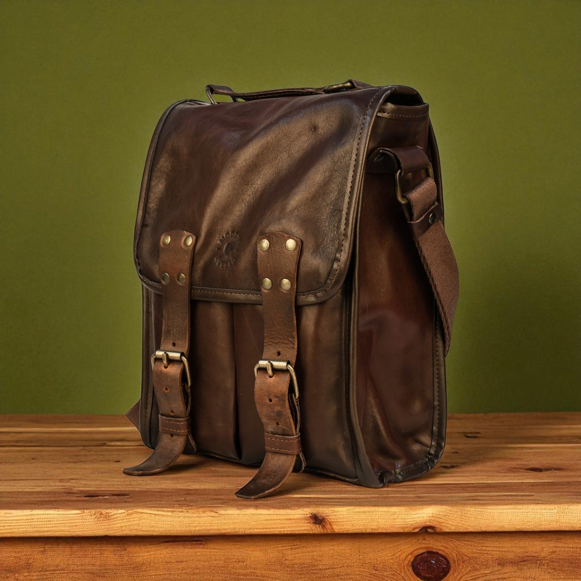 Brown bovine leather medium satchel bag with brass buckles & canvas strap on a wooden table, side view.