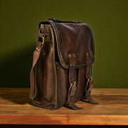 Brown bovine leather medium satchel bag with canvas strap on a wooden surface, showing front buckles and rivets.