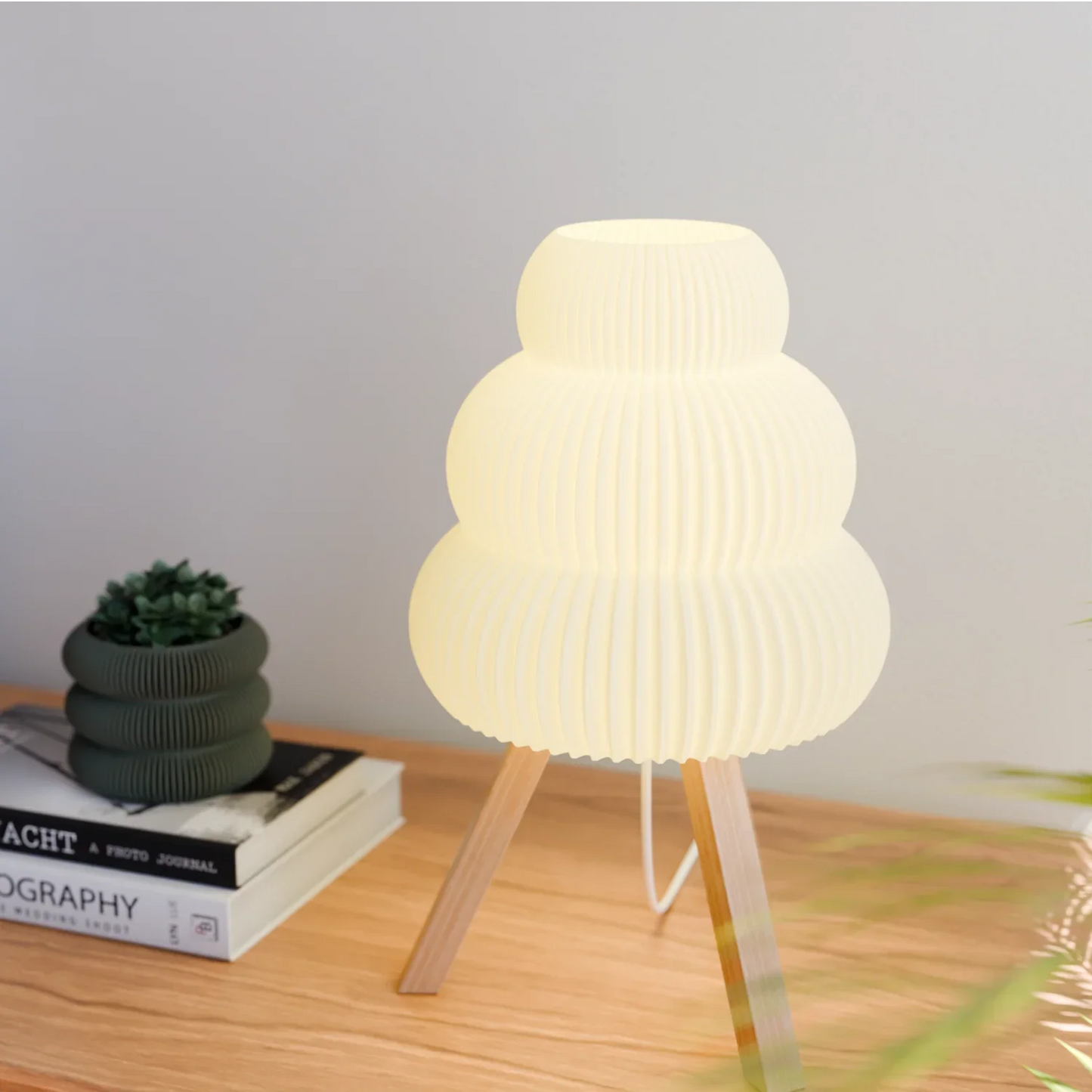 Modern tripod table lamp with white ribbed shade on wooden surface next to decorative plant and books
