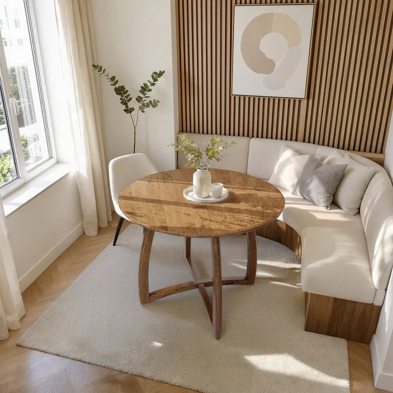 Modern dining nook with round wooden table, beige L-shaped bench, white chair, plant, and abstract wall art