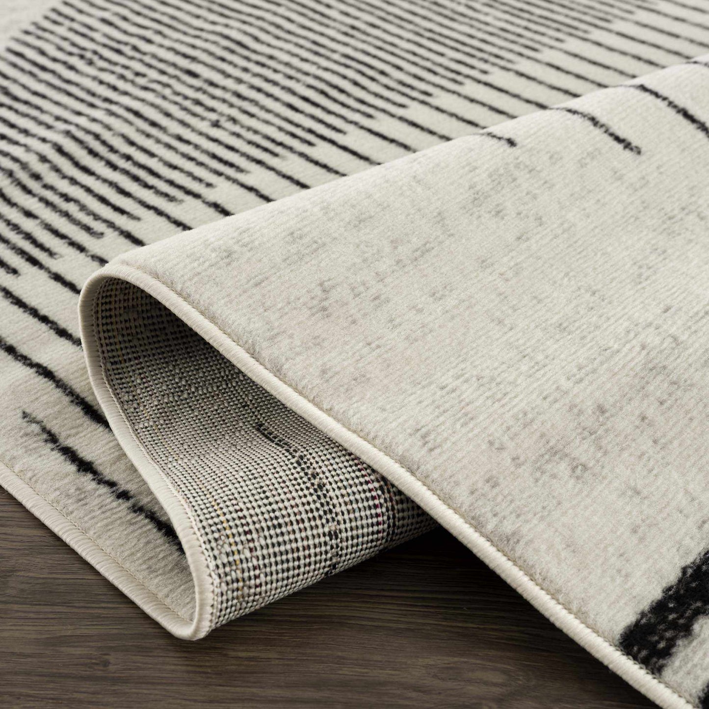 Close-up of Angus abstract black and white area rug, showing striped pattern, curled edge, and backing on wood floor.