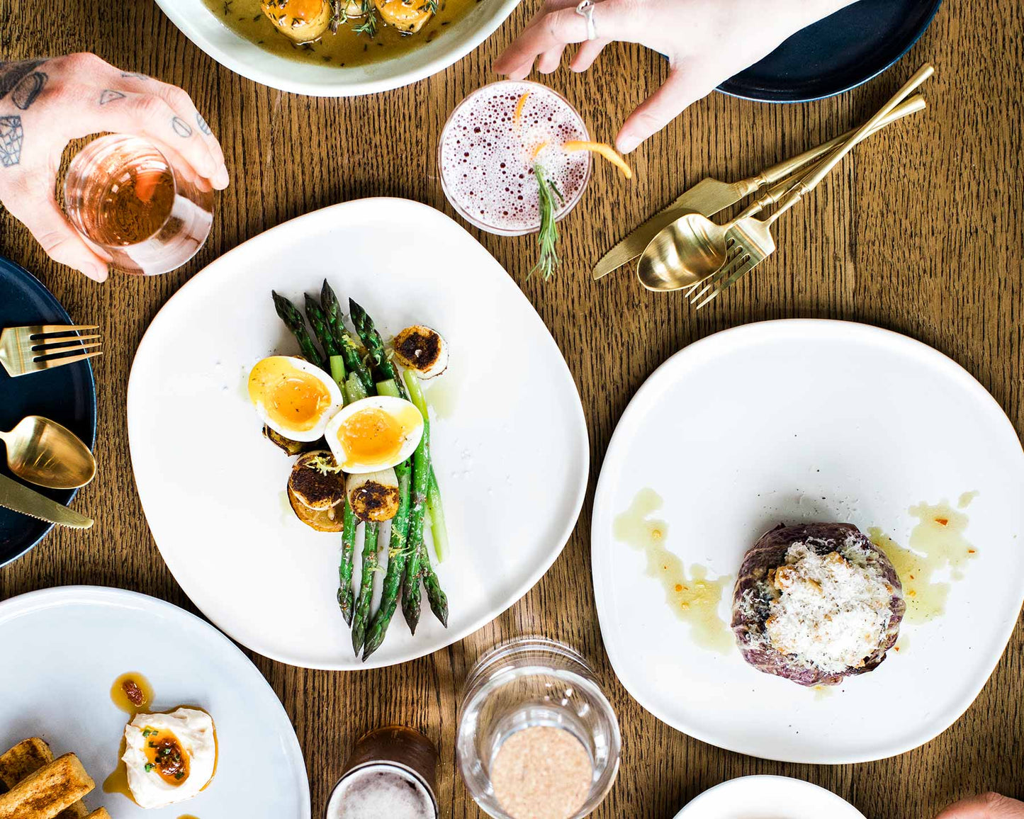 Gourmet dishes with asparagus, soft-boiled egg, roasted potatoes, and dessert on white plates with gold utensils on wooden table
