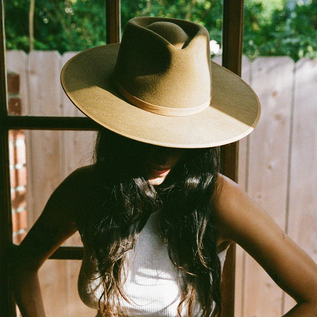 Woman wearing a wide-brimmed tan hat with long dark hair, standing indoors near a window