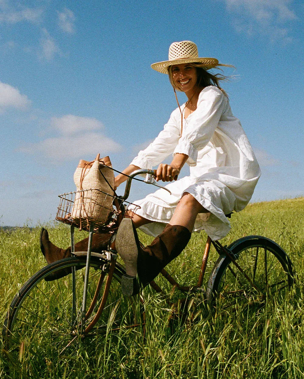 Smiling woman in white dress and straw hat riding vintage bicycle through green field under blue sky with woven bag in basket