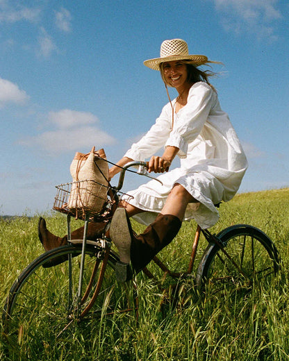 Smiling woman in white dress and straw hat riding vintage bicycle through green field under blue sky with woven bag in basket