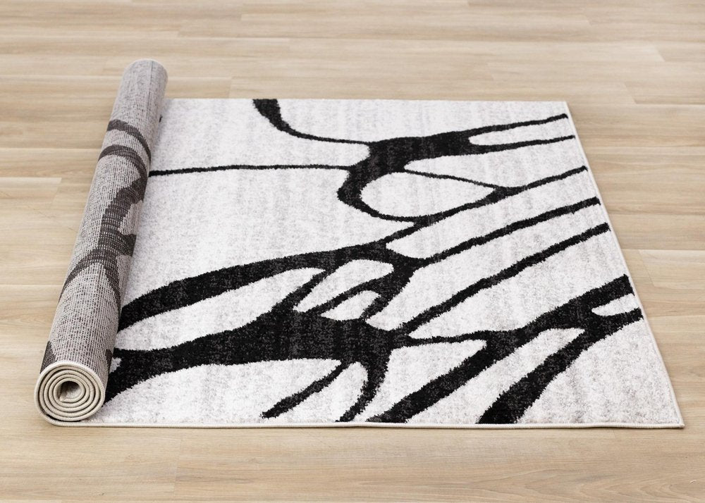 Black and gray abstract area rug partially rolled showing full linear pattern layout and jute backing on hardwood floor