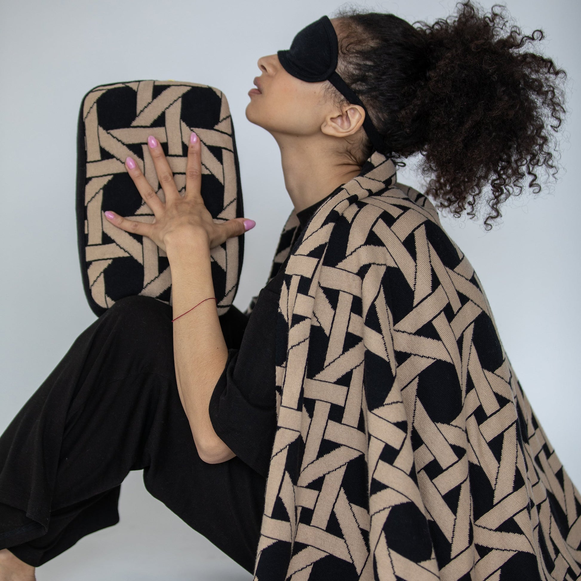 Woman wearing black geometric organic cotton travel blanket with eye mask holding matching compact pouch in dynamic pose