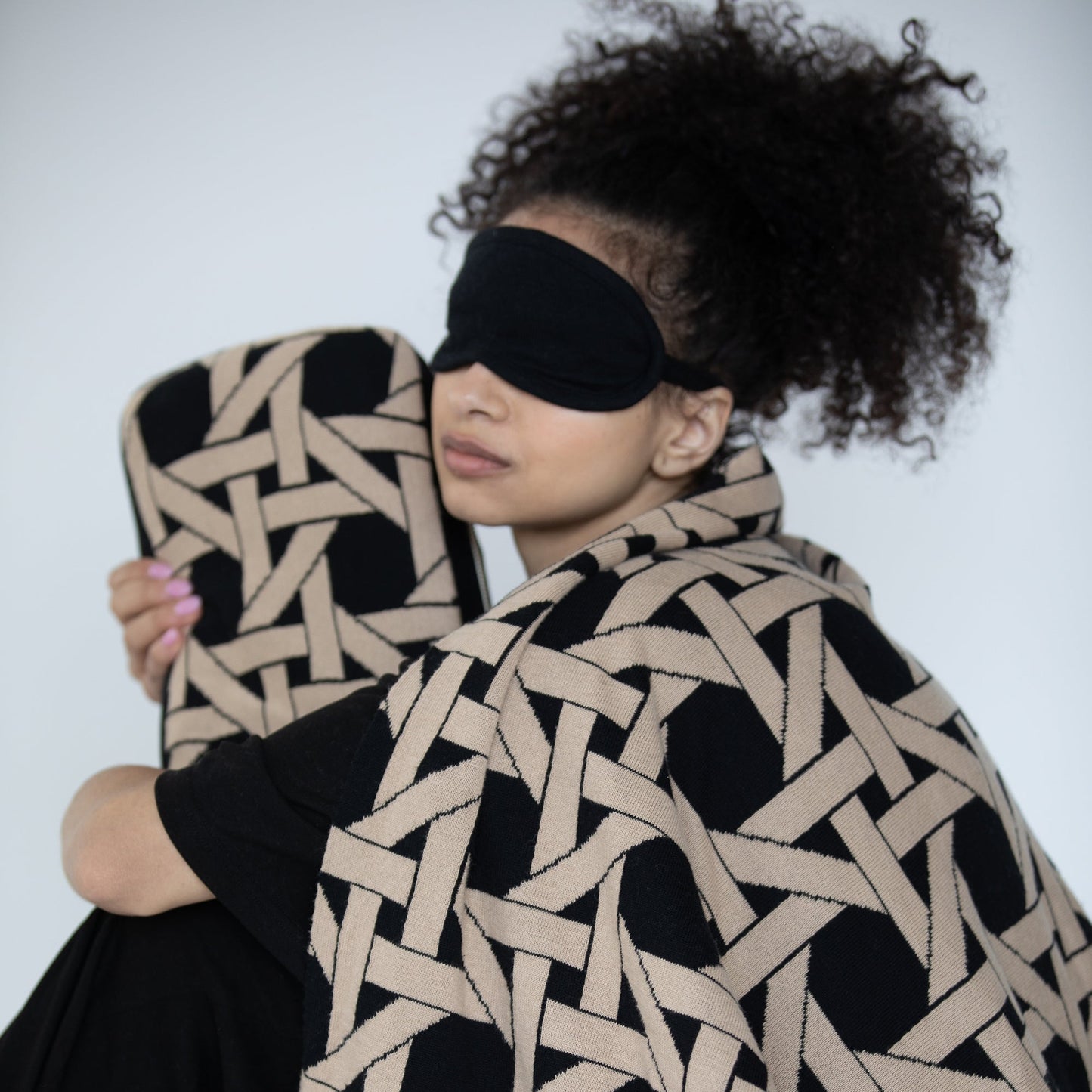 Woman wearing black organic cotton travel blanket draped over shoulders with matching eye mask and geometric compact pouch