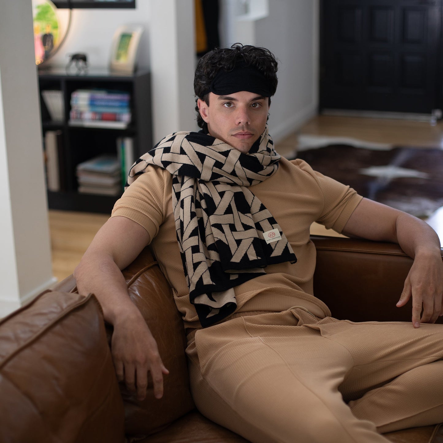 Man wearing black geometric organic cotton travel blanket draped as scarf with matching eye mask seated on leather sofa
