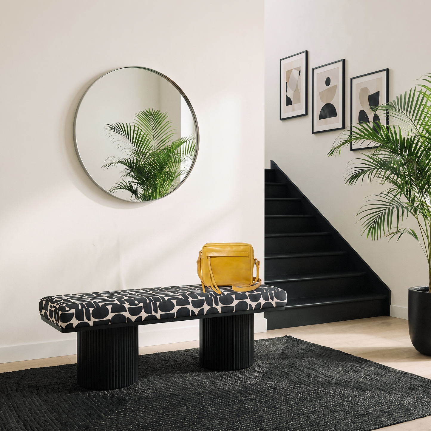 Black and white geometric upholstered bench with dual ribbed black pedestal legs styled in a modern entryway with round mirror black staircase and palm plant on dark area rug