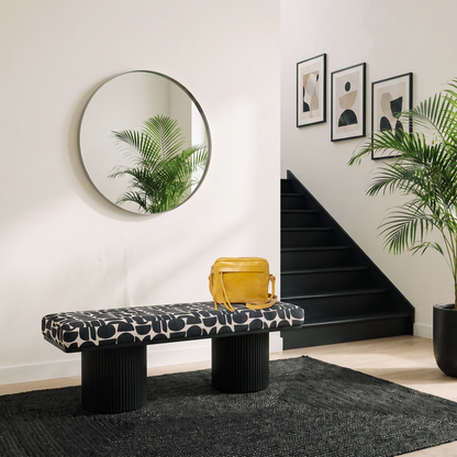 Black and white geometric upholstered bench with dual ribbed black pedestal legs styled in a modern entryway with round mirror black staircase and palm plant on dark area rug