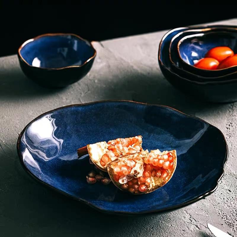 Lifestyle view of the Blue European Porcelain Dinnerware Set showcasing an unique egg shaped porcelain plate and matching dark blue glazed bowls, presented with fresh fruit on a stone surface.