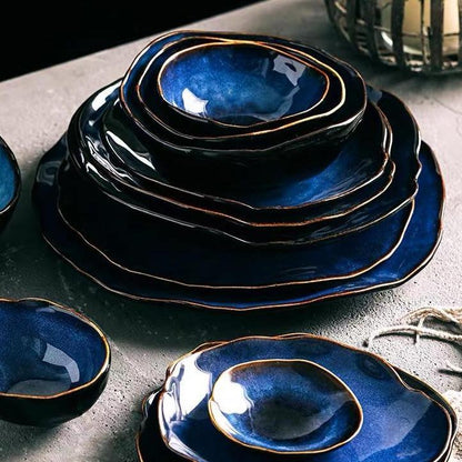 Blue European Porcelain Dinnerware Set from the Amelia Collection featuring dark blue glazed bowls and egg shaped porcelain plates with artisanal gold-trimmed edges on a textured surface.
