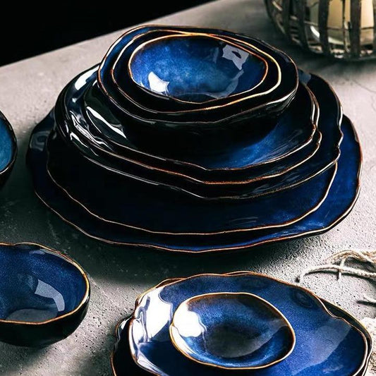 Blue European Porcelain Dinnerware Set from the Amelia Collection featuring dark blue glazed bowls and egg shaped porcelain plates with artisanal gold-trimmed edges on a textured surface.