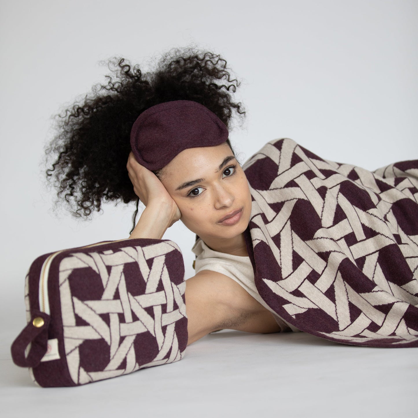 Woman resting with burgundy geometric organic cotton travel blanket and eye mask beside matching compact travel pouch