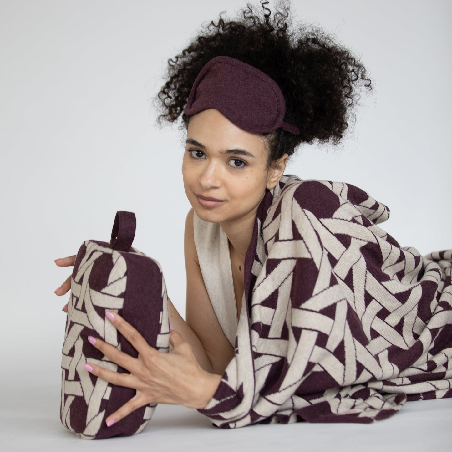 Woman wearing burgundy geometric organic cotton travel blanket draped over shoulders with matching eye mask and compact pouch