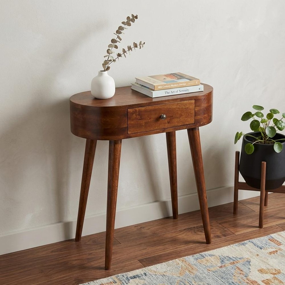Chestnut oval solid wood console table with drawer styled in entryway with white vase dried stems stacked books and potted plant on hardwood floor