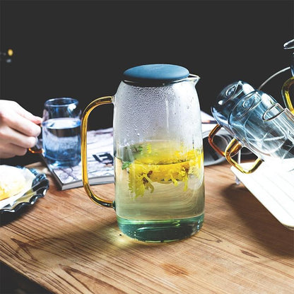 Clear glass carafe with gold handle and blue tinted base filled with yellow flowers on wooden table