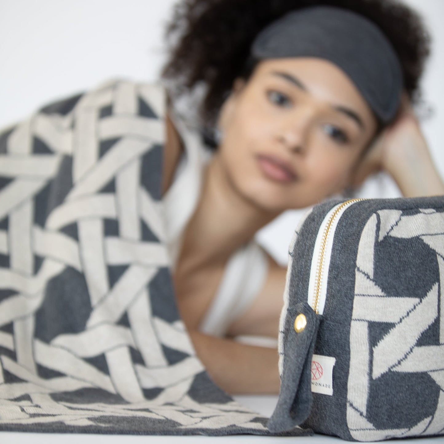Woman resting with dark gray geometric organic cotton travel blanket and matching eye mask beside coordinating compact pouch