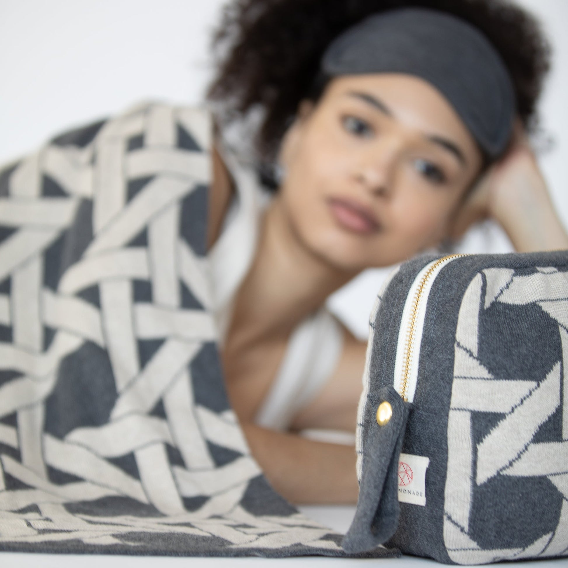 Woman resting with dark gray geometric organic cotton travel blanket and matching eye mask beside coordinating compact pouch