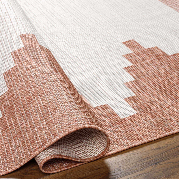 Close-up of Hauteloom Djugun Peach Fuzz indoor/outdoor rug unrolling on a wood floor, showing Aztec geometric pattern